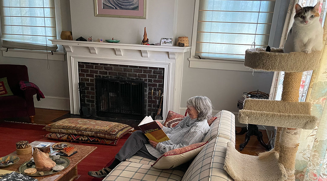 A photo of professor Virginia Johnson sitting on her couch and reading a math proofs book with her cat by the window.
