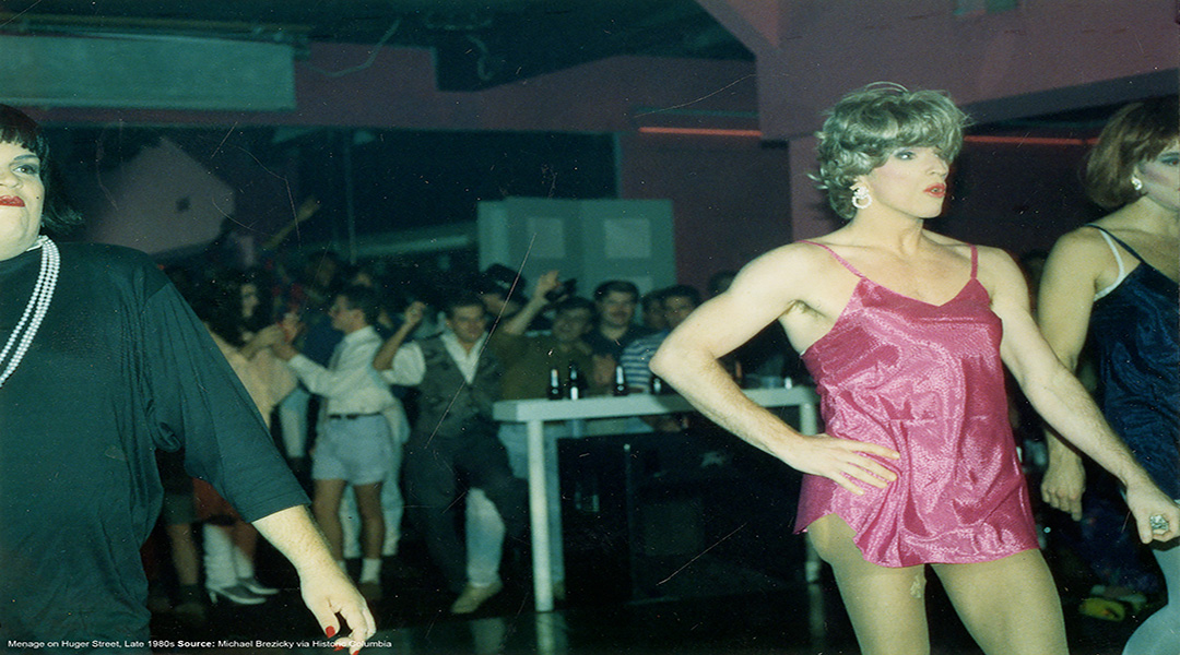 a person in drag participates in a pageant at menage with the crowded dance floor in the background
