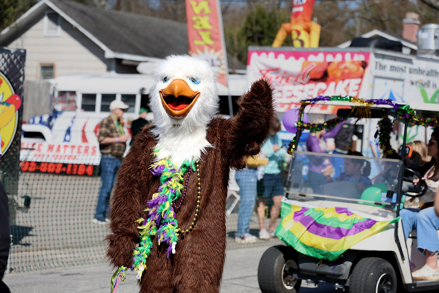 Mardi Gras_HC_1038cc_small A person dressed in a large eagle mascot costume waves while wearing Mardi Gras beads in purple, green, and gold. Behind them, a decorated golf cart and parade attendees line the street during a Mardi Gras parade.