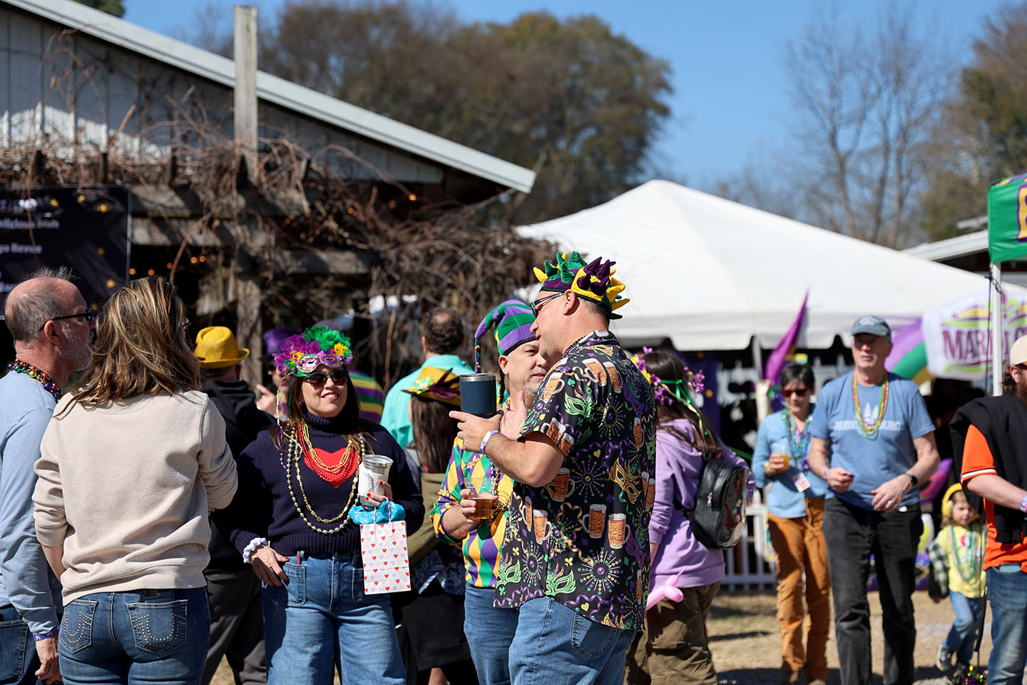 Mardi Gras_HC_1073cc_small A group of people wearing Mardi Gras beads and festive hats in purple, green, and gold stand outdoors talking and holding drinks. Tents are visible in the background during a Mardi Gras celebration.