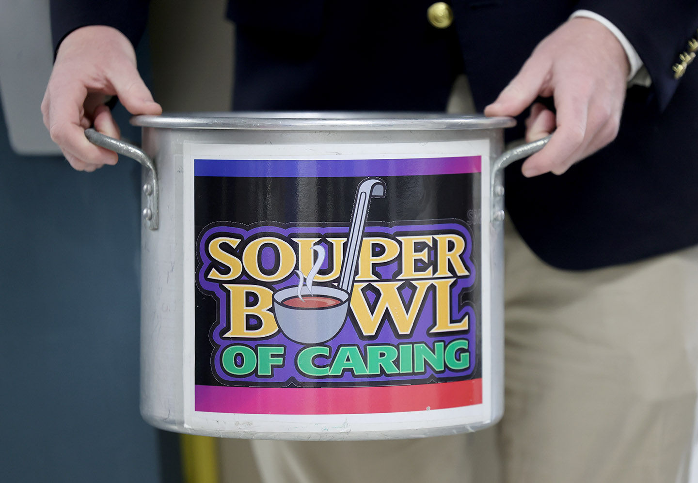 SouperBowl_HC_0478cc_small A student in a suit holds a pot labeled "Souper Bowl of Caring" outside the doors. His hands are holding the handles of the pot. He is wearing a navy suit with gold buttons and khaki pants.