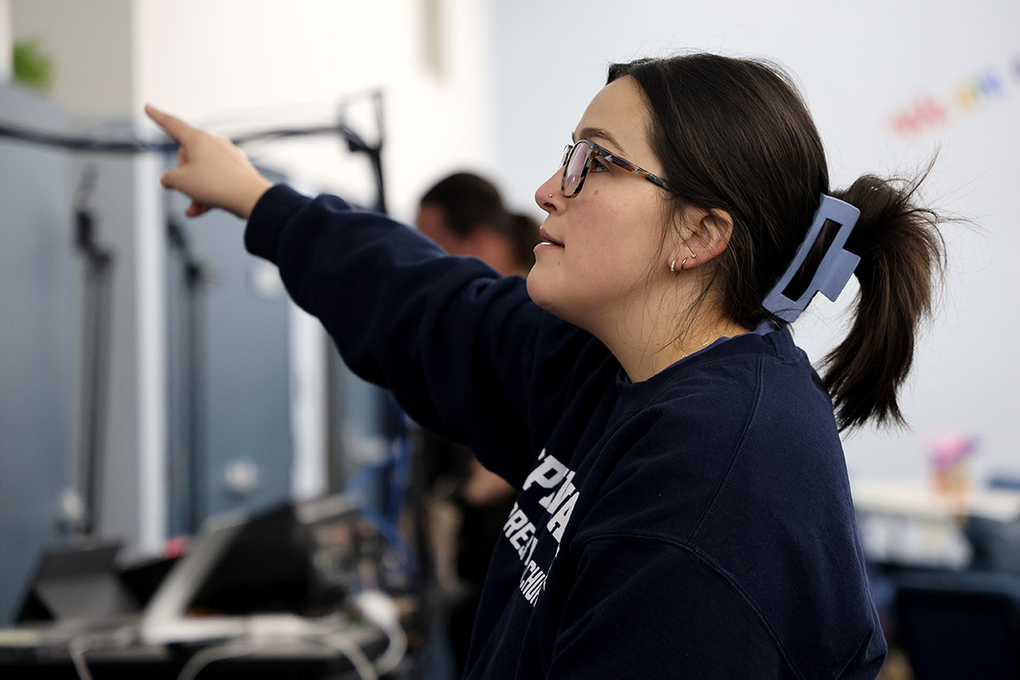 Meredith Orel points to the left of the image. She can be seen looking off into the distance. She is wearing a navy blue sweatshirt, glasses, and has her hair in a ponytail with a blue claw clip. 