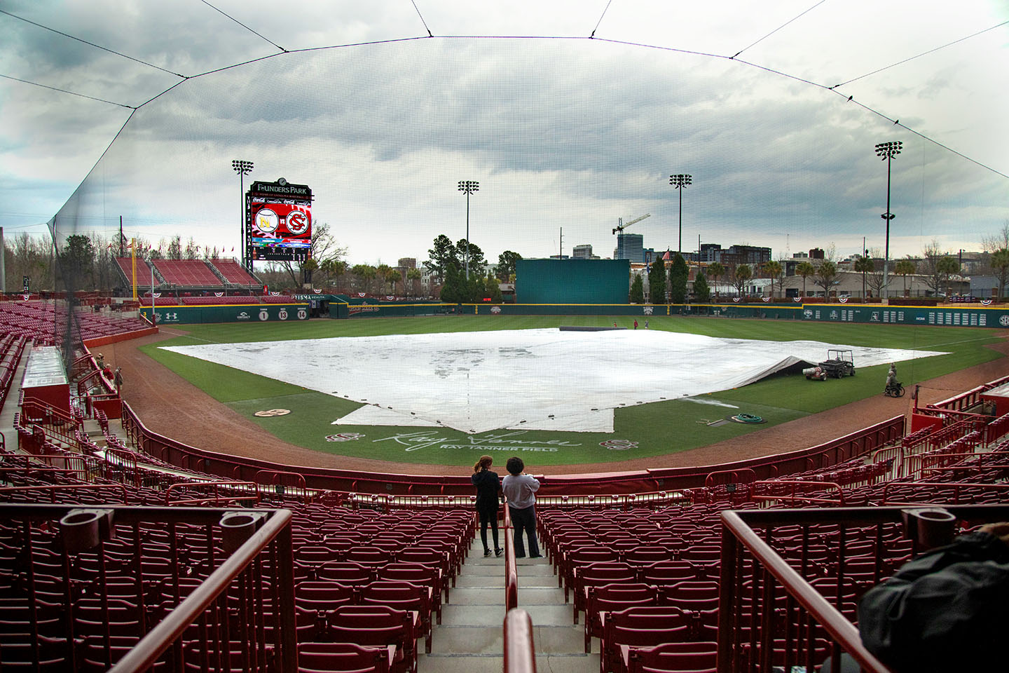 Empty baseball seats look out over a tarp that covers the baseball field while storm clouds loom in the distance.