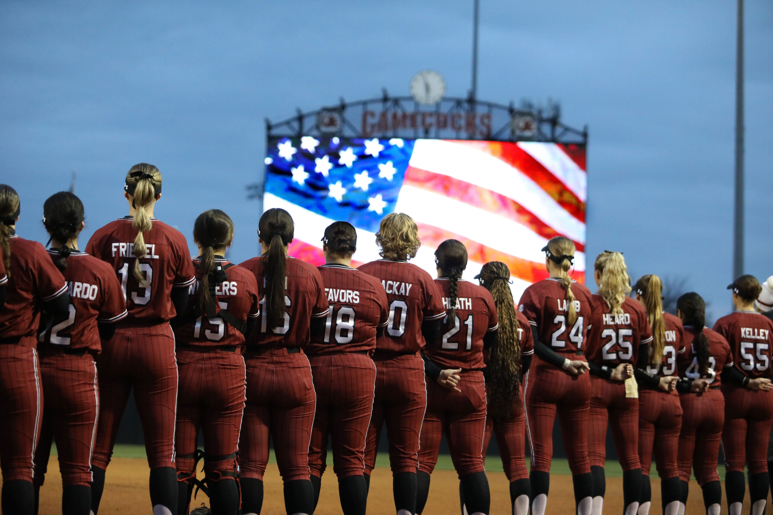 Players form a straight line as seen from behind as they stand at attention for the National Anthem. In the background is a projection of the American flag. 