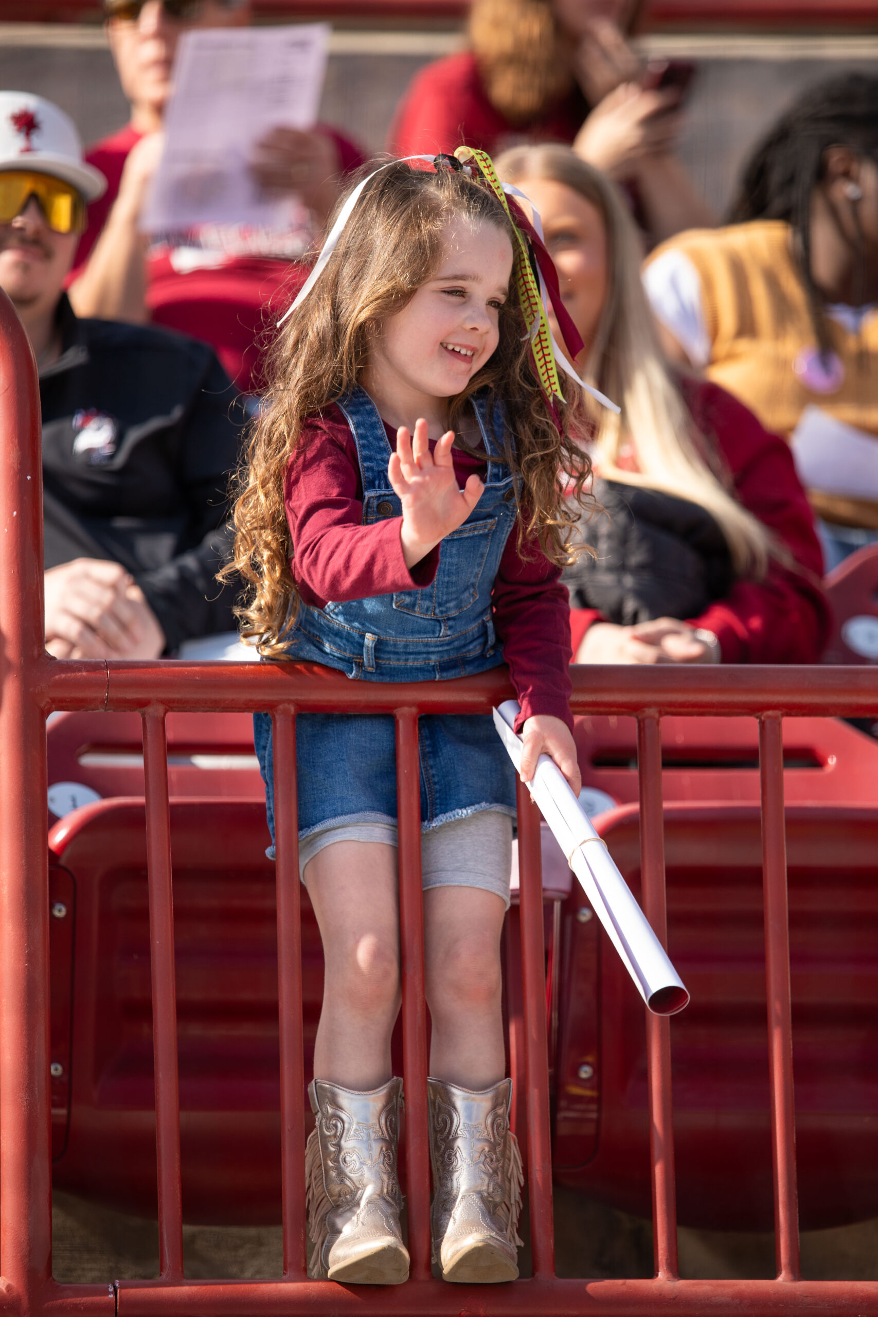A little girl in jean shorts and a garnet top waves expectantly from the stands. 
